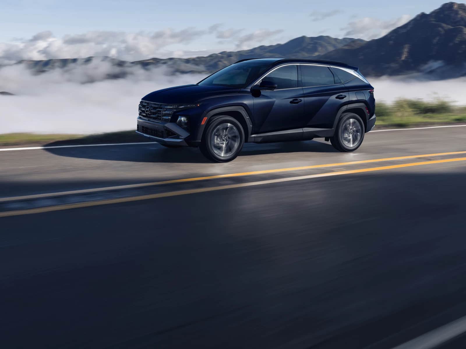 Hyundai Tucson 2026 bleu foncé sur une route de montagne, mer de nuages en contrebas, ciel bleu
