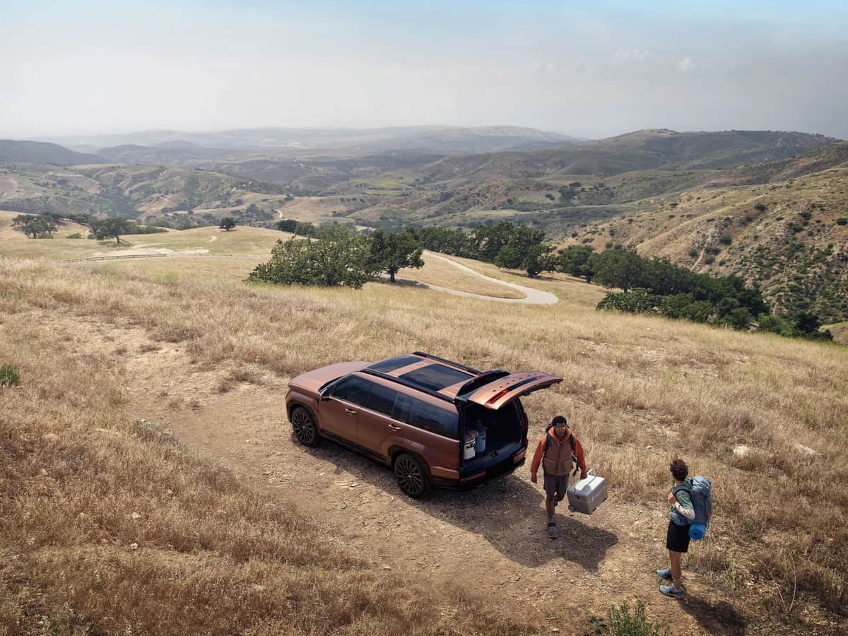 Hyundai Santa Fe 2026 cuivré, hayon ouvert, deux randonneurs qui déchargent leur équipement dans un champ vallonné