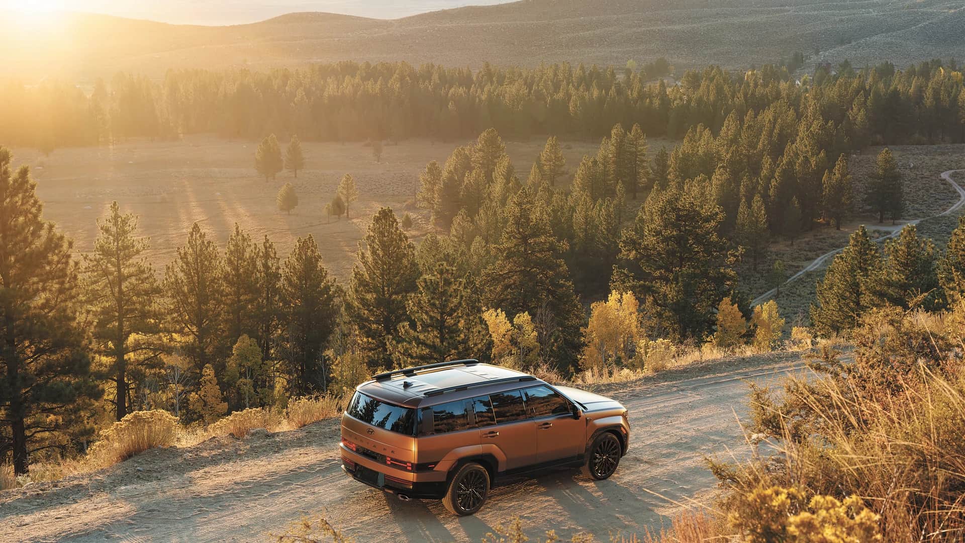 Vue aérienne d'un Hyundai Santa Fe 2026 sur un chemin de terre, forêt de conifères et vallée dorée au soleil couchant