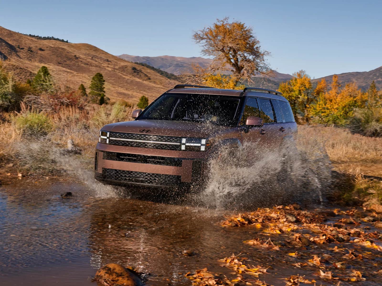 Hyundai Santa Fe 2026 cuivré qui traverse un ruisseau dans un paysage d'automne, feuillage orangé et collines