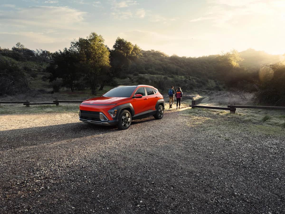 Hyundai KONA 2026 orange garé au départ d'un sentier, deux randonneurs, lumière de fin de journée
