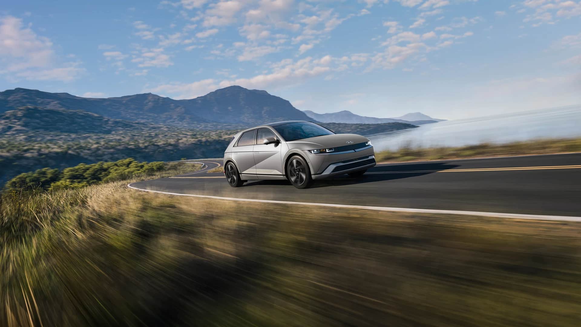 Hyundai IONIQ 5 2026 argent sur une route en lacets qui longe la côte, montagnes et ciel bleu