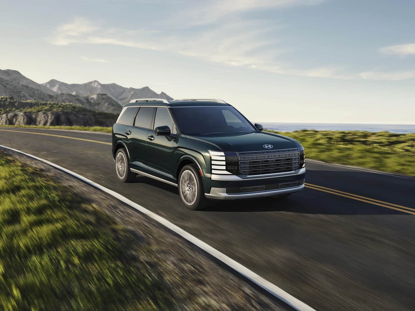 Hyundai Palisade 2026 vert foncé, trois quarts avant, sur une route de campagne bordée de prés, montagnes au loin